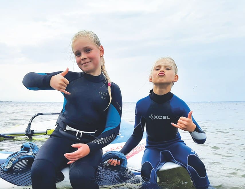 Zwei Kinder im Neoprenanzug auf einem Surfbrett im Bodden
